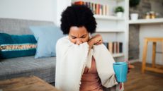 Woman can be seen sat on a wooden floor coughing into her elbow while she holds a cup of tea in a blue mug in the other hand. The background is blurred, however, she is sat next to a wooden coffee table and a grey couch with blue cushions. There is also a bookshelf behind her and a wooden stool against a kitchen counter to the right