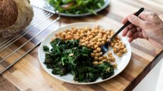 An old hand holding a fork, eating a plate of leafy green vegetables and chickpeas